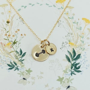 May include: Gold necklace with two charms. One charm is a dumbbell and the other charm is a letter K.