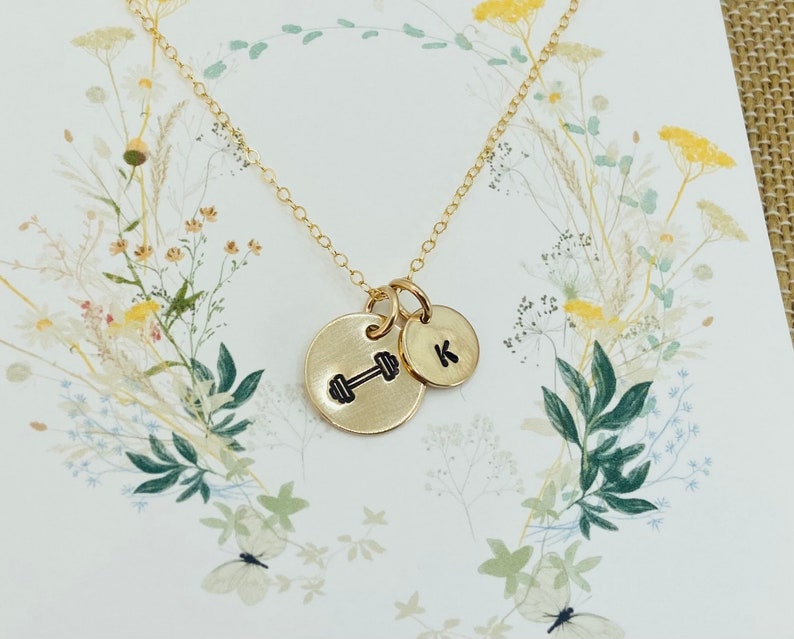 May include: Gold necklace with two charms. One charm is a dumbbell and the other charm is a letter K.