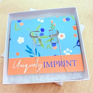 May include: A pair of silver heart-shaped drop earrings displayed in a white box. The earrings are on a blue card with floral designs and the text "Uniquely IMPRINT". The box sits on a tan surface.