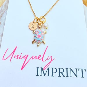 May include: A gold-colored necklace featuring a turtle pendant with pink and blue enamel detailing. The necklace includes a small initial charm and a pearl. The word "Uniquely" is written in pink script, and the word "IMPRINT" is in navy blue.