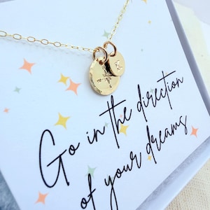 May include: A gold-colored necklace with two pendants. One pendant is a round disc with a compass design, and the other is a smaller disc with the letter "K". The card behind the necklace reads "Go in the direction of your dreams."