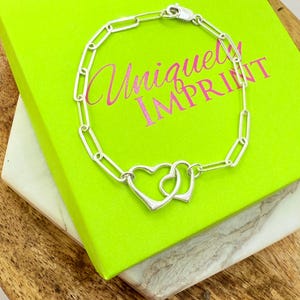May include: A silver bracelet with a chain link design and two connected heart charms. The bracelet rests on a bright green box with the text "Uniquely Imprint" in pink. The box sits on a wooden surface.