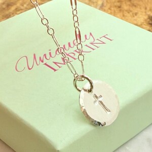 May include: A silver necklace with a small, round pendant featuring a cross design. The pendant hangs from a delicate chain. The necklace is displayed on a light green box with the text "Uniquely INPRINT".