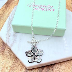 May include: A silver-toned necklace with a flower pendant. The pendant is a detailed, five-petal flower with a mother-of-pearl effect. The necklace has a toggle clasp and is displayed on a marble surface with a mint green box that reads "Uniquely Mprint".