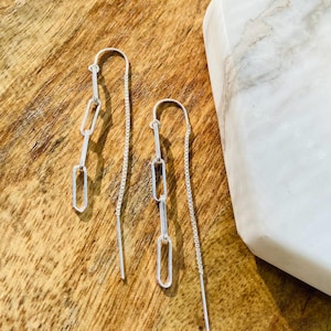 May include: A pair of silver dangle earrings with a chain and three rectangular links. The earrings are on a wooden surface.