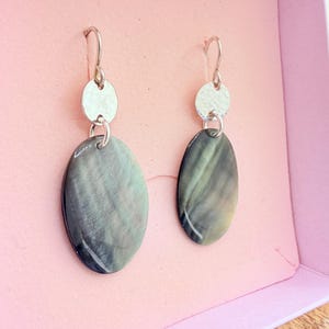 May include: A pair of dangle earrings with silver-tone hooks and circular accents. Each earring features an oval, iridescent, gray-green stone. The earrings are displayed in a pink box.