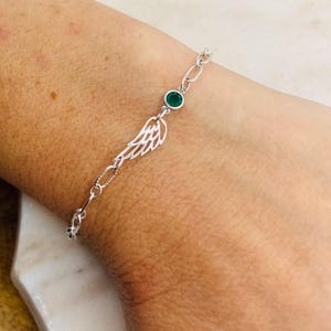 Puede incluir: Un brazalete de cadena de plata con una piedra preciosa verde y un delicado encanto de ala de plata.