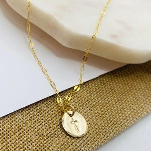 May include: A gold chain necklace with a small, round pendant featuring a hammered texture and a cross design.