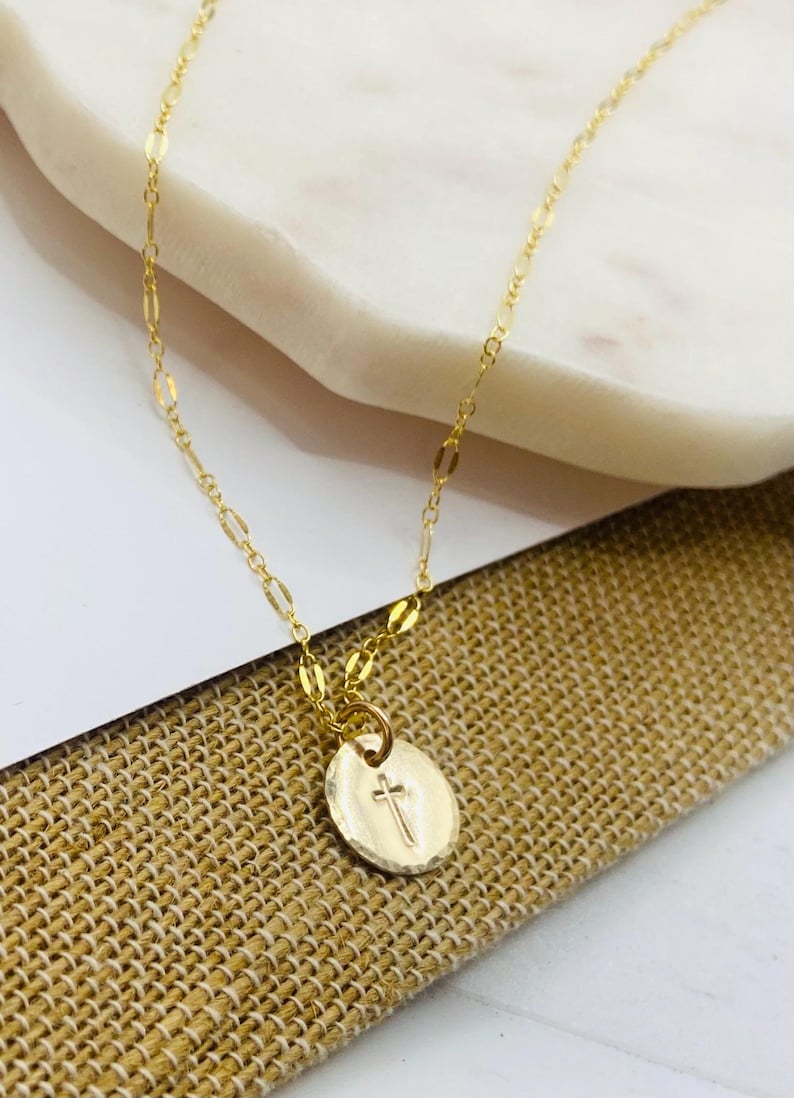 May include: A gold chain necklace with a small, round pendant featuring a hammered texture and a cross design.