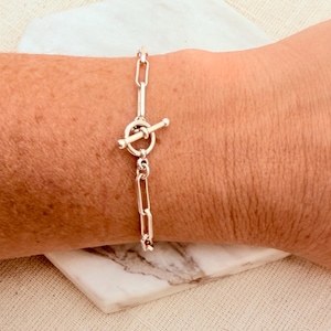 May include: A silver-toned chain bracelet featuring elongated rectangular links and a circular toggle clasp closure. The bracelet is secured with a T-bar that passes through the ring, creating a polished and metallic accessory for the wrist.