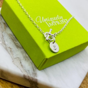 May include: A silver necklace with an oval pendant featuring the letter "E" rests on a bright green box. The box is labeled "Uniquely Imprint" in cursive. The necklace is displayed on a white and gray marble surface.