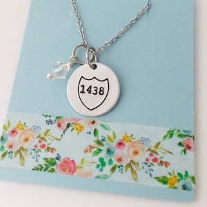 May include: A silver necklace with a round pendant featuring the number 1438 inside a shield shape. A small clear crystal charm hangs from the pendant.