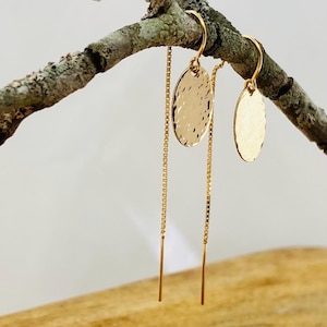 May include: Gold-coloured dangle earrings. Each earring has a textured, oval-shaped charm suspended from a delicate chain. The earrings are displayed on a natural, textured branch against a soft background.