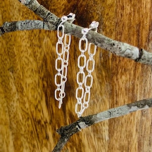 May include: Silver chain earrings with an elongated oval link design. The earrings hang from a small stud and are displayed against a natural wood background. The earrings are a simple, yet elegant accessory.