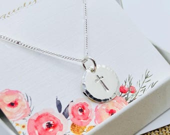 May include: A silver necklace with a delicate chain and a round pendant featuring a small cross. The pendant is textured and has a hammered finish. The necklace is displayed in a white box with a floral design.