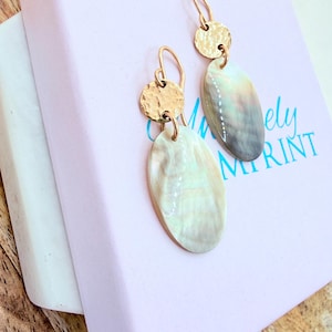 May include: Gold-tone earrings with a hammered disc and an oval, iridescent shell-like pendant. The earrings are displayed on a light pink box with the text "lovely imprint" in teal.