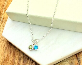 May include: A silver necklace with two round pendants, one green and one blue. The necklace is displayed on a white surface with the text "Uniquely IMPRINT" on a box in the background. The background is a wooden surface.