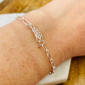 May include: A silver chain bracelet with a delicate angel wing charm. The bracelet is on a person's wrist.