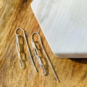 May include: A pair of silver threader earrings featuring a delicate chain that transitions into a series of three elongated, rectangular paperclip-style links. The earrings are constructed from polished silver metal, showcasing a slender, minimalist design that hangs vertically from a curved ear wire.