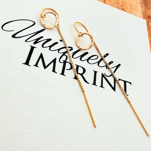 May include: Gold-toned threader earrings with a small circle design. The earrings are displayed on a white surface with the words "Unique Imprint" in black script.