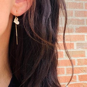 Puede incluir: Un colgante de corazón de oro cuelga de una delicada cadena de oro, que cuelga de la oreja de una persona.