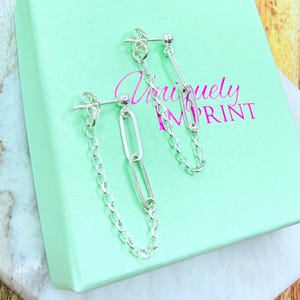 May include: A pair of silver chain earrings with a chain and oval link design. The earrings are displayed on a light green box with the text "Uniquely Imprint" in pink script.