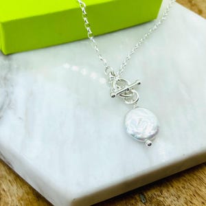 May include: A silver necklace with a large, white, coin-shaped pearl pendant. The necklace has a toggle clasp and a delicate chain. The necklace is displayed on a white marble surface, with a green box in the background.