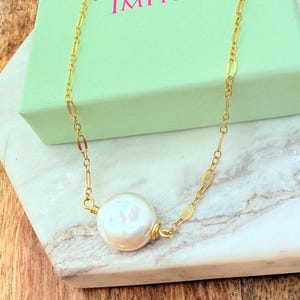 May include: A gold-colored chain necklace with a large, iridescent pearl pendant. The necklace is displayed on a marble surface next to a light green box with the text "Uniquely Imprint".