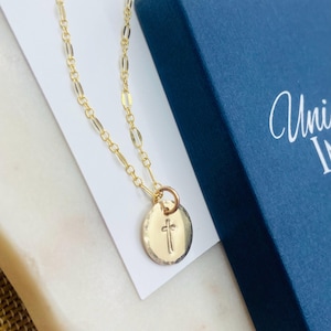 May include: A gold chain necklace with a small round pendant featuring a cross design. The necklace is displayed on a white surface with a blue box in the background.