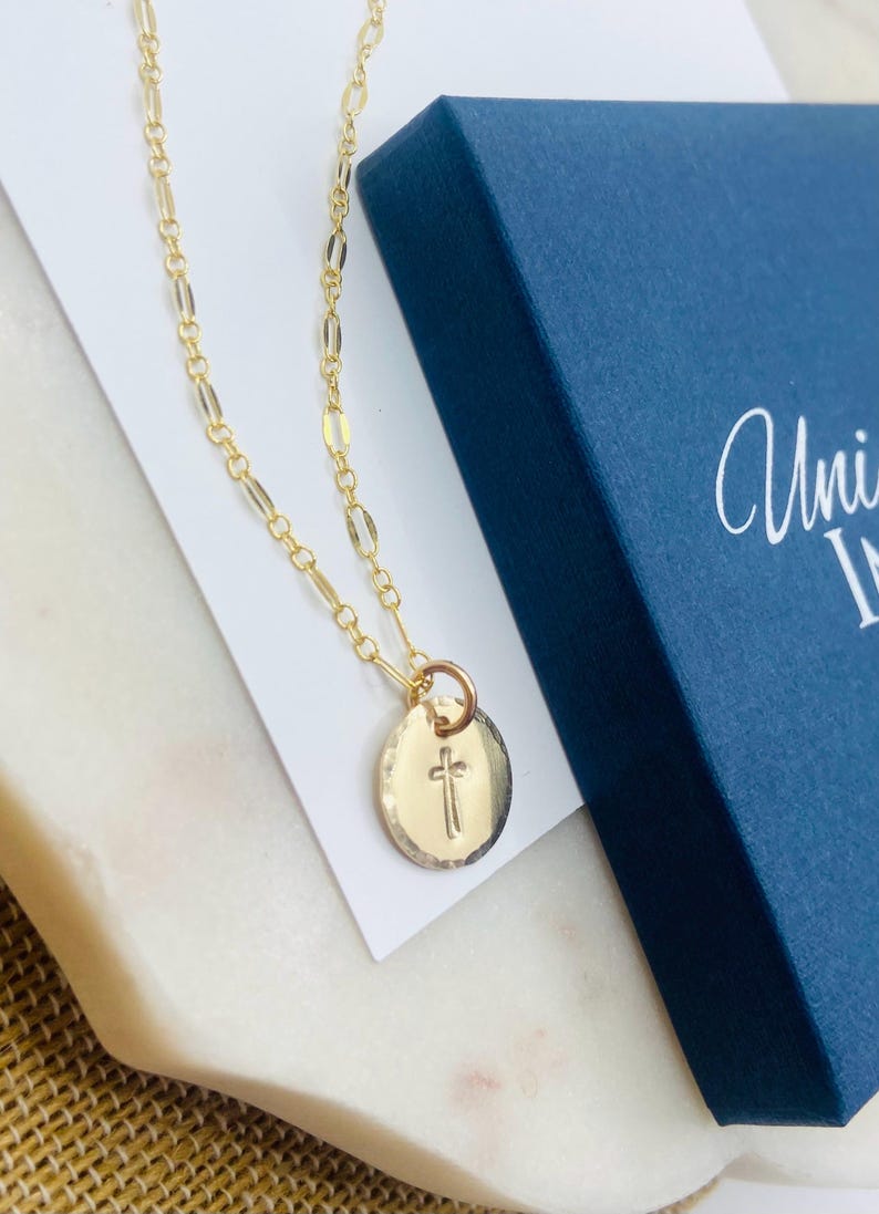 May include: A gold chain necklace with a small round pendant featuring a cross design. The necklace is displayed on a white surface with a blue box in the background.