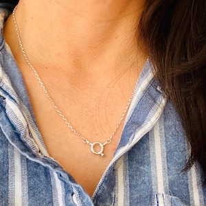 May include: A silver chain necklace featuring a round spring ring clasp positioned at the front. The necklace is made of thin, polished silver links and rests against a blue and white vertically striped collared shirt with a button-down front.