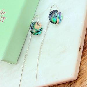 May include: A pair of abalone shell earrings with a silver chain and hook. The earrings have a round, iridescent abalone shell disc with shades of green, blue, and purple. The earrings are displayed on a white marble surface.