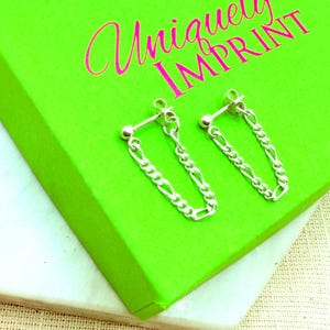 May include: A pair of silver stud earrings featuring a dangling figaro chain design. Each earring consists of a small spherical stud connected to both ends of a short silver chain, creating a looped effect. The text Uniquely IMPRINT is visible.