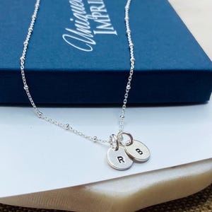 May include: A silver necklace with a delicate chain and two small round charms with the letters "R" and "B" engraved on them. The necklace is displayed on a white surface with a blue box in the background.