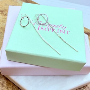 May include: A pair of silver drop earrings with a hammered circle design and a delicate chain. The earrings are displayed on a light green box with the text "Uniquely Imprint". The box is stacked on a pink box and a white marble surface.