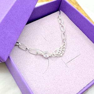 May include: A silver-toned necklace with a wing design and a clear, round gemstone is displayed inside a purple gift box. The necklace has a chain-link style and is presented on a light pink velvet-like material.