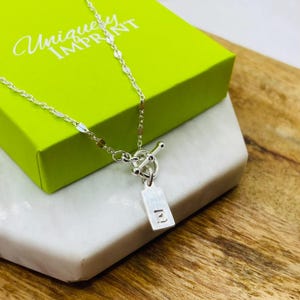 Peut inclure: Un collier en argent avec un pendentif rectangulaire portant la lettre "E". Le collier a un fermoir à bascule et est présenté sur une surface en marbre blanc, avec une boîte verte en arrière-plan qui indique "Unique Imprint".