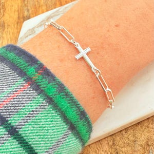 May include: A silver-colored bracelet with a cross pendant. The bracelet features a chain-link design and is worn on a wrist. The cross is centered on the bracelet. The person is wearing a green, blue, and red plaid shirt.
