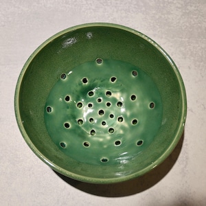 May include: A round, green ceramic bowl featuring a series of small, evenly spaced drainage holes across the bottom. The interior surface has a glossy, smooth finish with a slightly darker green hue, while the outer rim is a lighter matte green.