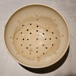 May include: A round ceramic colander with a cream-colored glaze and a speckled finish. The interior features numerous drainage holes distributed across the base and sides, with subtle brown and tan tonal variations in the glaze texture.