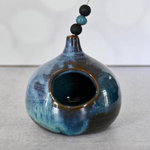 May include: A ceramic birdhouse with a blue and brown glaze. The birdhouse has a round body with a large opening and a small neck. Three decorative beads hang from a wire above the birdhouse.