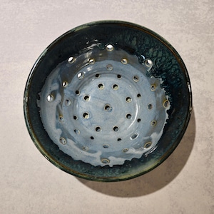 May include: A round ceramic colander bowl featuring a deep blue and light grey glazed finish. The interior surface is marked by a series of evenly spaced circular drainage holes arranged in a radial pattern, extending from the center to the rim.