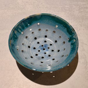 May include: A ceramic berry bowl featuring a glazed finish with a deep teal rim that transitions into a light blue interior. The bowl is perforated with numerous circular drainage holes evenly spaced across the bottom and sides of the vessel.