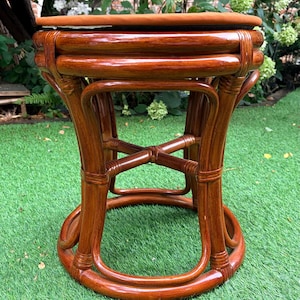 May include: A round stool constructed from polished brown rattan with a smooth, glossy finish. The base features a circular frame connected by curved vertical supports and a central X-shaped brace. A flat, circular, burnt-orange cushion sits on top.