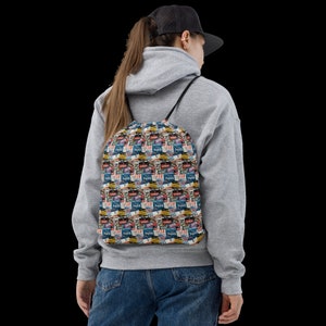 May include: A white drawstring bag with a colourful pattern of vintage travel stickers. The bag has a black drawstring and is being worn by a person.