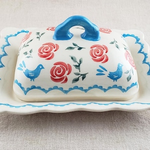 May include: A white ceramic butter dish with a lid, decorated with red roses, blue birds, and green leaves. The lid has a blue handle. The dish has a scalloped edge with a blue trim.