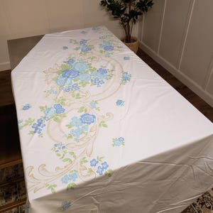 May include: White tablecloth with a floral design featuring blue and light blue roses and green leaves. The floral pattern is accented with gold scrollwork. The tablecloth is rectangular and covers a table.