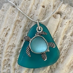 Può includere: Una collana con un ciondolo in vetro di mare turchese e un ciondolo a forma di tartaruga argentato. Il ciondolo ha la forma di una lacrima e ha una superficie liscia e lucida.