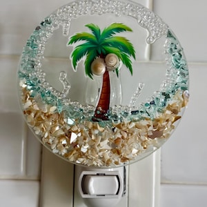May include: A white nightlight with a round glass shade featuring a palm tree and seashells. The shade is decorated with small pieces of broken glass and seashells.