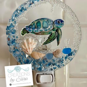 May include: A decorative night light featuring a sea turtle design. The night light has a clear, circular frame with blue glass pieces and seashells. The night light has a white base with the text "Creations by Sara".